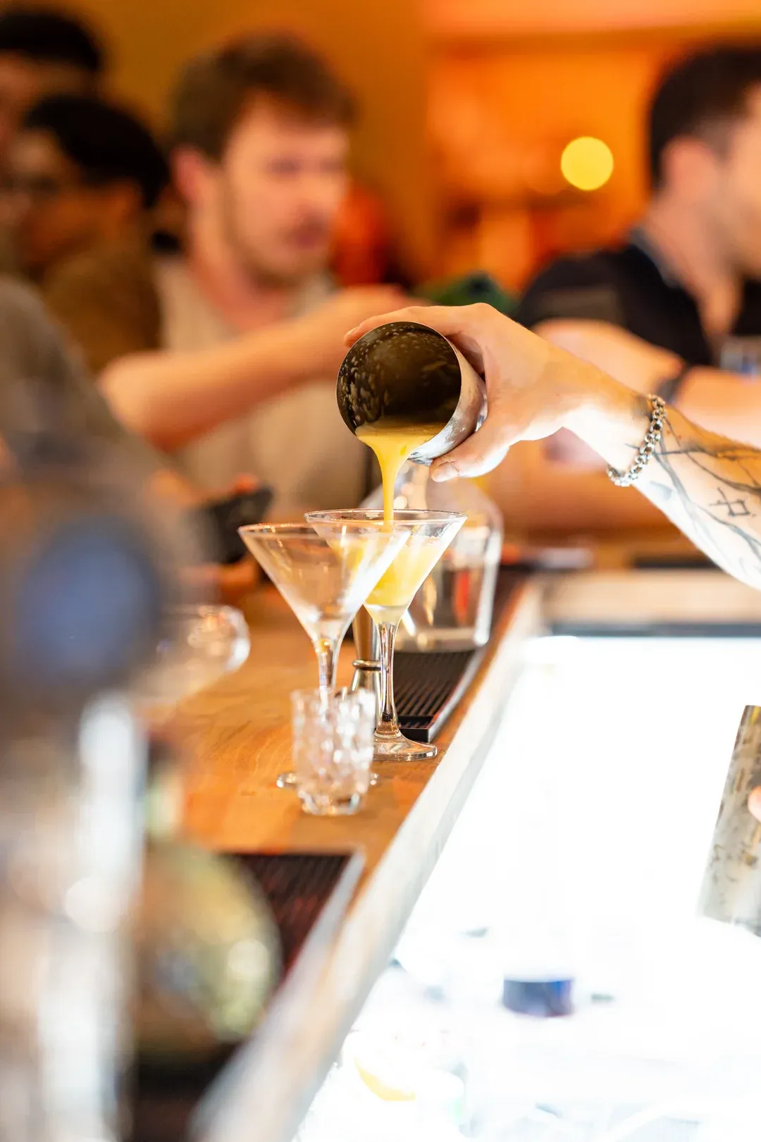 A yellow cocktail poured from the shaker into a martini glass, the crowd blurred along the bar