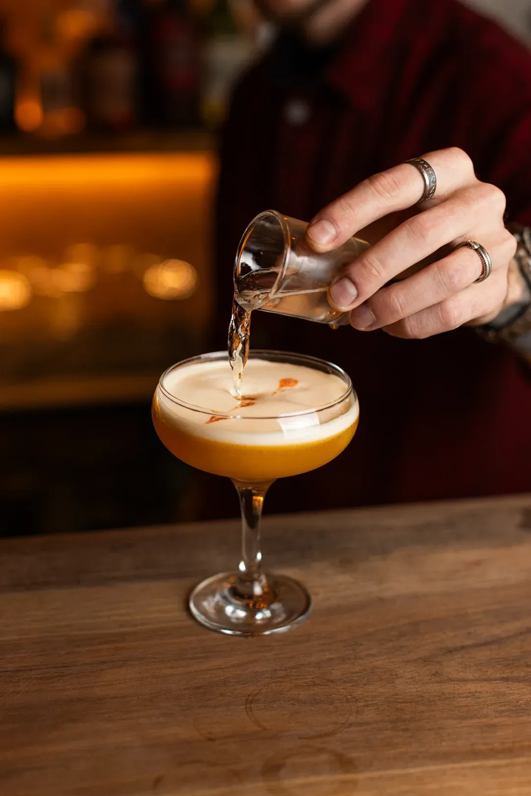 A bartender pouring a shot into a foam-topped cocktail at the bar counter